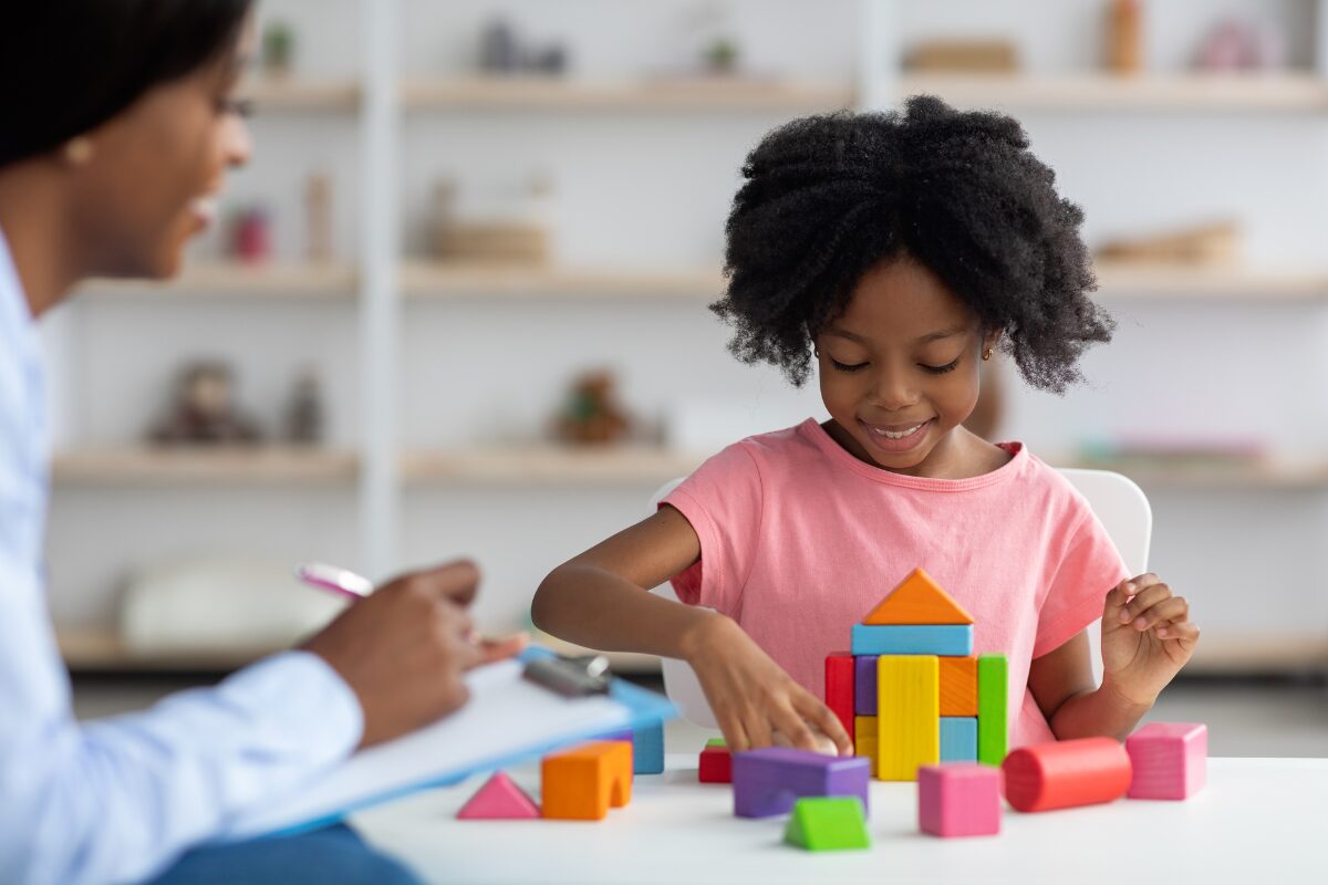 A young child plays with building blocks next to a therapist as part of play-based therapy.