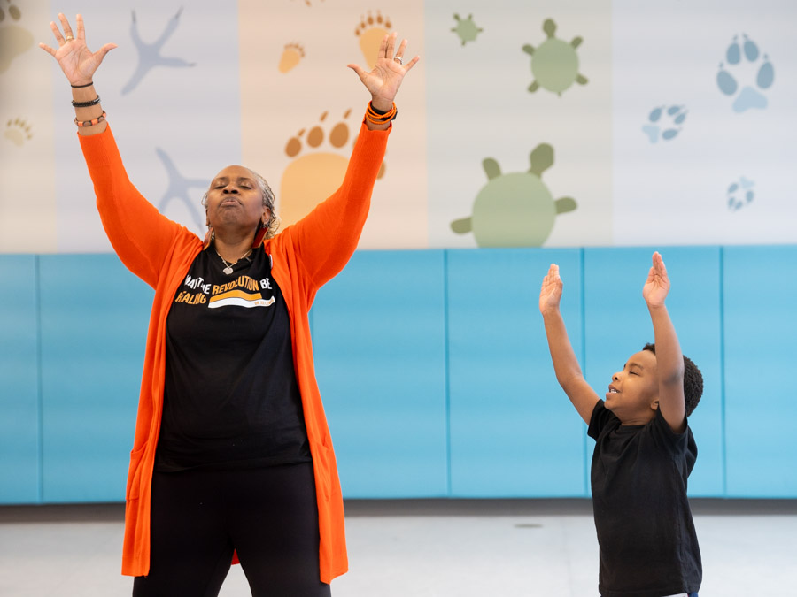 Preschool facilitator guiding a young child through a mindfulness activity, both stretching calmly and focusing on their breathing in a classroom setting.